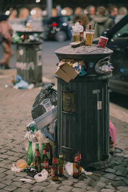 A black, cylindrical public rubbish bin with a domed lid is situated on a cobbled pavement in an urban setting. The bin's surface shows a matte finish and has visible signs of use, with various paper documents and small cardboard pieces attached to its exterior, along with a partially open flap revealing the contents inside. On top of the bin, there are several disposable coffee cups with plastic lids, some with straws, and a small paper food container, indicating they are used for takeaway beverages and food. Surrounding the base of the bin, there are numerous empty glass and plastic bottles, including beer and soft drink bottles, some lying on their sides, and crumpled paper and tissues scattered on the ground. In the background, blurred figures of pedestrians and parked cars, along with additional litter, suggest a busy street environment, possibly in a commercial or public area. The scene reflects typical urban waste accumulation that services like rubbish removal companies manage, emphasizing the importance of effective disposal to maintain cleanliness. The lighting is natural, indicating daytime, with overcast skies that diffuse the sunlight evenly across the scene.