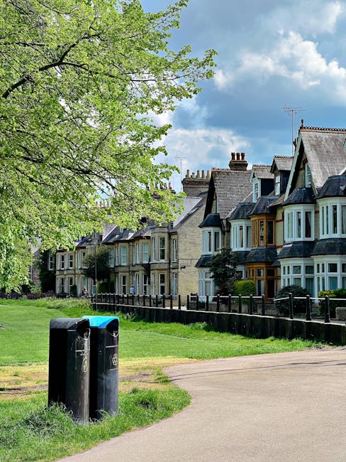 The image shows a row of traditional Victorian-style terraced houses with bay windows and slate roofs, situated along a residential street. In the foreground, there are two black outdoor mailboxes positioned next to a light-colored paved pathway, which leads through a grassy, well-maintained communal area. On the left side of the image, a large tree with fresh green leaves extends over the scene, providing shade and a natural backdrop against a partly cloudy sky with patches of blue. The houses appear tidy and well-kept, with small front gardens and metal railings separating them from the pathway. The environmental setting suggests a peaceful, suburban neighbourhood, and the presence of the mailboxes and pathway indicates common areas that could potentially be involved in community maintenance or private waste collection arrangements, such as those managed by local waste disposal services like rubbishcollectionkingstonuponthames.co.uk, which may handle alternative waste or on-site clearance for these residential properties.