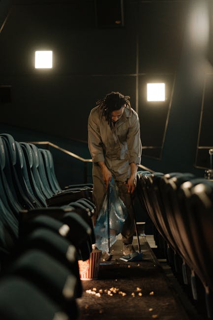 A person with dark, curly hair and wearing a grey jumpsuit is in a dimly lit auditorium, bending down to pick up or clear discarded popcorn from the dark, carpeted floor between rows of dark green or grey plastic seats. The seats are arranged in an orderly fashion on either side of the central aisle and have a smooth, matte finish with armrests. The background features dark walls with two illuminated square lights providing minimal ambient lighting, creating a moody atmosphere. The individual appears to be engaged in cleaning or waste collection within the auditorium, which could relate to independent rubbish removal services or on-site clearance in a public or private entertainment venue, aligned with the themes of rubbish and waste management services often associated with venue maintenance or event cleanup. The scene subtly reflects scenarios where prompt and professional waste handling is needed after a public event, supporting the importance of efficient rubbish collection and disposal.