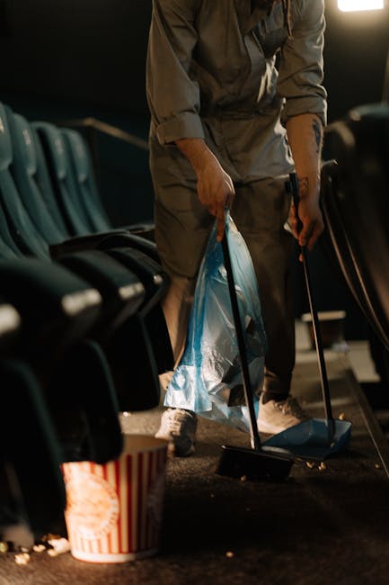 A person dressed in casual, neutral-coloured clothing is seen standing amidst a collection of empty blue and black plastic chairs positioned in a staggered arrangement on a dark indoor floor. They are using a dustpan and brush to sweep up debris, which includes small scattered pieces of rubbish such as paper, plastic, and other waste items. The individual holds a bright blue bin bag in one hand, ready for collection, and appears to be engaged in clearing the area. Nearby, a striped red and white popcorn-style bucket is placed on the floor, partially filled with additional litter. The environment suggests a public venue, possibly an auditorium or theatre space, with a dimly lit background illuminated by artificial lighting. The scene illustrates a process of on-site waste clearance, consistent with private rubbish disposal or cleanup services often employed for event waste management, as provided by companies like rubbishcollectionkingstonuponthames.co.uk, to maintain cleanliness and hygienic standards in such spaces.