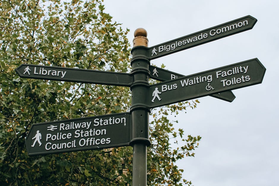 A black, multi-directional street signpost standing outdoors against a backdrop of green leafy trees and an overcast sky. The signs display white lettering and symbols indicating directions to various locations, including Biggleswade Common at the top, followed by Library, Bus Waiting Facility, Toilets, Railway Station, Police Station, and Council Offices. Each sign has a walking figure icon, with the Bus Waiting Facility sign featuring an additional wheelchair accessible symbol. The pole has a rounded finial at the top and the signs are attached with metal clamps. The scene suggests an urban or town centre setting, where clear directional guidance is provided for pedestrians and visitors. Such signage could be relevant in discussions about local amenities or alternative ways of managing waste and waste removal on communal or public property, reflecting the organized approach typical in civic environments overseen by local councils, similar to Kingston upon Thames.