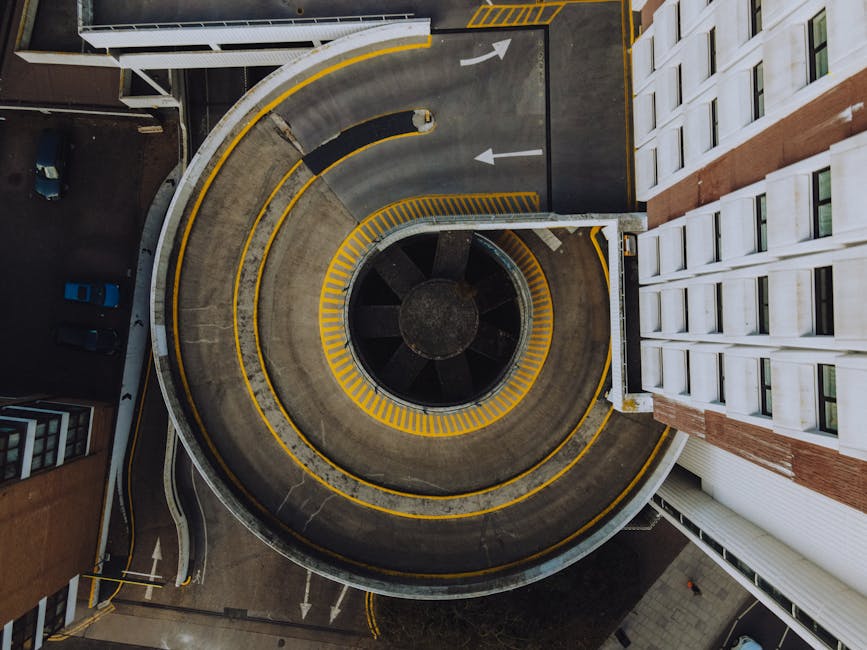 An aerial view of a multi-level spiral parking structure with concrete surfaces, featuring yellow safety markings along the edges and ramp outlines. The circular ramp curves around a central cylindrical core, which is dark and weathered. Surrounding the parking facility, there are adjacent buildings, including a modern white high-rise with numerous rectangular windows on the right side, and a smaller brown-brick building on the lower left. The environment appears urban, with cars parked on the lower levels and a dark asphalt driveway with white directional arrows guiding vehicle flow. The scene is lit with natural daylight, emphasizing the textures of concrete and the contrast of yellow safety lines. The image subtly connects to waste management themes by highlighting structures possibly associated with commercial or residential waste handling areas within an urban estate, suggesting potential access points for skip or rubbish collection services like those offered by Kingston-based providers.