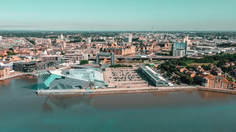 An aerial view of a modern urban waterfront area showing a large, angular building with a metallic, reflective surface and a light blue-green hue, situated on the riverbank. The building's facade features geometric panels and a combination of smooth and textured finishes, with a nearby parking lot containing several cars. Behind it, there are multiple levels of a parking structure, and further back, a densely built cityscape with a mix of historic brick buildings and contemporary high-rises. The scene is illuminated with natural daylight, with the river in the foreground reflecting parts of the structures, and expansive city skyline with a clear sky extending into the distance. The image conveys an environment where private property and modern development coexist near water, characteristic of areas that could involve independent waste management or rubbish removal services in urban settings, as supported by visual cues of construction and infrastructure development around the waterfront.