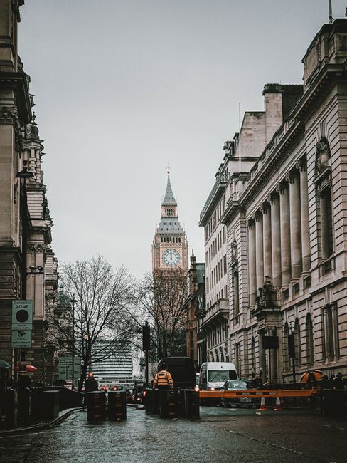A city street scene featuring the historic clock tower commonly known as Big Ben in London, visible in the background with its pointed spire and detailed clock face. The foreground shows a wet asphalt road with several vehicles, including a black van and a small white truck, parked or moving along the street. On the street, there are orange safety barriers and traffic cones indicating some form of on-site clearance or temporary access control. Tall, classical-style buildings with ornate stone facades and columns line both sides of the street, their surfaces showing signs of weathering and detailed architectural ornamentation. Leafless trees are visible along the pavement, suggesting a late autumn or winter setting. The overall atmosphere appears overcast, with diffused natural light illuminating the scene, supporting a neutral and professional tone. This urban environment relates to alternative waste handling or rubbish removal services, with the scene illustrating typical city logistics for maintaining clear public spaces or conducting private clearance activities, as might be managed by local or independent providers like rubbishcollectionkingstonuponthames.co.uk.