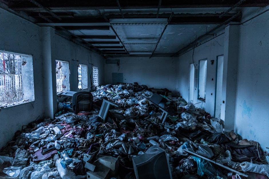 The image shows a large, cluttered indoor space with a high ceiling and multiple windows along one wall, allowing natural light to partially illuminate the scene. The floor is completely covered with a mixture of discarded items, primarily consisting of black and clear plastic rubbish bags, some torn open revealing contents such as paper, cardboard, and miscellaneous household debris. Scattered among the waste are broken electronic devices, including old computer monitors and a computer chair, whose dark fabric and plastic components are partially obscured by the trash. The walls of the room are painted in a light color but appear stained and worn, with visible patches of peeling paint and grime. The ceiling frames are exposed, showing metal beams and ductwork, which give an industrial feel to the environment. Outside the windows, faint outlines of trees and fencing can be seen, indicating the space might be a vacant building or storage area awaiting clearance. This scene reflects a visual example of an extensive rubbish accumulation suitable for professional waste removal services, such as those offered by rubbishcollectionkingstonuponthames.co.uk, highlighting the need for independent or commercial clearance in private or neglected properties.