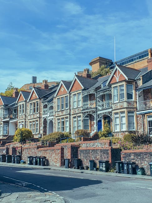 A row of Victorian-style terraced houses with ornate white-painted ironwork on the balconies and window frames, featuring brick facades and pitched roofs with gables, situated on a slightly elevated street with a brick retaining wall and a line of black and green wheelie bins at the curb. The houses are set against a bright blue sky with some sparse clouds, and a modern building with glass windows and metal structures is visible behind the rooftops. The environment suggests an urban residential area where private or independent waste collection may be arranged, reflecting aspects of alternative rubbish handling and clearance services such as those provided by companies like rubbishcollectionkingstonuponthames.co.uk. The scene emphasizes residential waste management, with no visible activity occurring in the image.