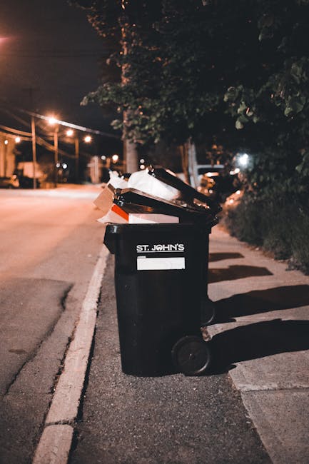 A black plastic waste bin with the label 'ST. JOHN'S' placed on a suburban sidewalk at night, filled with various discarded items including flattened cardboard boxes and plastic wrappers. The bin is positioned near the curb, casting shadows on the pavement under streetlights. In the background, a row of streetlights illuminates a quiet residential street with trees, parked cars, and houses visible in the distance. The scene reflects an instance of private rubbish disposal or on-site waste collection, with the bin awaiting collection or removal, which relates to non-local-authority rubbish management services. The overall atmosphere is calm and dimly lit, emphasizing the bin's role in waste management in a typical urban or suburban setting.