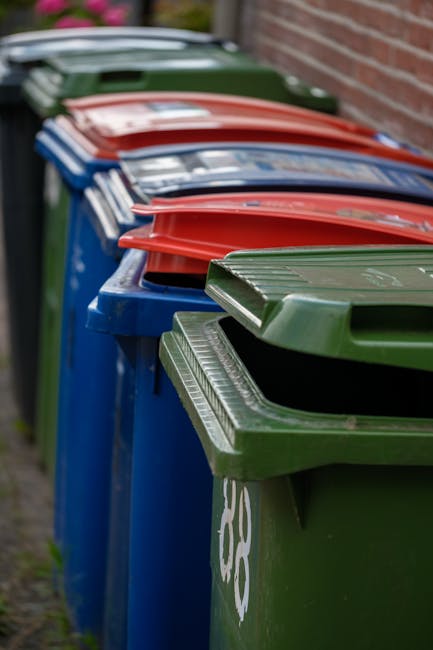 A black plastic waste bin with the label 'ST. JOHN'S' placed on a suburban sidewalk at night, filled with various discarded items including flattened cardboard boxes and plastic wrappers. The bin is positioned near the curb, casting shadows on the pavement under streetlights. In the background, a row of streetlights illuminates a quiet residential street with trees, parked cars, and houses visible in the distance. The scene reflects an instance of private rubbish disposal or on-site waste collection, with the bin awaiting collection or removal, which relates to non-local-authority rubbish management services. The overall atmosphere is calm and dimly lit, emphasizing the bin's role in waste management in a typical urban or suburban setting.