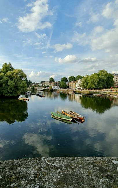 A tranquil riverside scene showing a calm waterway reflecting a partly cloudy blue sky. Several small boats, with textures of weathered wood and painted surfaces in shades of green, orange, and brown, are moored along the riverbank in the foreground. On the far side of the river, there are leafy green trees and a variety of residential buildings and houses, some with white or pale facades, situated along the shoreline. The environment suggests a peaceful, shaded area suitable for private or independent boat mooring and occasional riverbank clearance. The overall atmosphere is bright and natural, with light casting gentle reflections on the water surface, and the scene points to a typical outdoor location where local waste management services, such as those provided by rubbishcollectionkingstonuponthames.co.uk, may assist with waste disposal after outdoor activities or maintenance in the area.