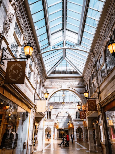 The image depicts an indoor shopping arcade with a high glass roof supported by ornate metal framework, allowing natural light to illuminate the space. The architecture features classical stone facades with decorative moldings, large arched openings, and elaborate cornices. On either side of the central walkway, there are retail storefronts with display windows, some showcasing clothing and merchandise, while others have signage. Warm-glow wall-mounted lanterns with metal fixtures hang along the corridor, providing additional ambient lighting. The polished stone floor is smooth and reflective, with a few shoppers walking or browsing, and a person pushing a shopping cart is visible in the distance. The environment appears clean and well-maintained, indicative of a traditional shopping mall or historic shopping arcade, where private management might handle waste disposal through independent rubbish collection services, supporting the need for thorough rubbish removal solutions within such commercial spaces. The scene emphasizes a structured, elegant interior setting associated with the type of environment that benefits from professional waste management to maintain its aesthetic appeal and cleanliness.