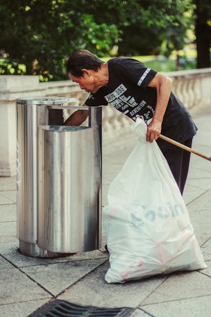 A middle-aged man with dark hair, dressed in a black t-shirt with white text and dark trousers, is seen leaning forward to dispose of a white plastic bag filled with rubbish into a cylindrical, stainless steel bin with an open top. The bin is situated on a paved sidewalk in an outdoor urban setting, with a stone balustrade and lush green trees in the background. The man is holding a wooden stick in his left hand, which supports keeping the bin lid open, and his right hand is placing the bag inside. The scene is illuminated by natural daylight, highlighting the metallic finish of the bin and the textured pavement. The image reflects an act of community-based or independent waste disposal, aligning with rubbish removal services offering on-site clearance or private rubbish collection, such as those provided by companies like rubbishcollectionkingstonuponthames.co.uk.