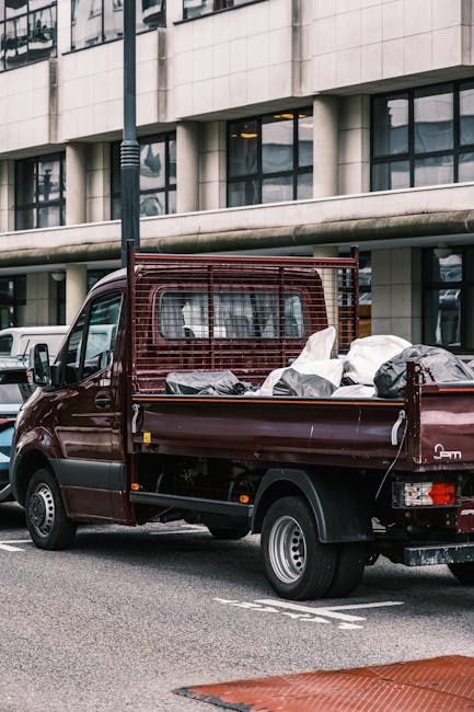 A small, dark red flatbed truck parked on an urban street in front of a modern, multi-storey office building with large, rectangular windows and a light-colored facade. The truck's open cargo area is filled with an assortment of waste materials wrapped in white plastic and black garbage bags, indicating it is engaged in private waste or rubbish removal. The vehicle features a metal safety grille at the cab's rear, and the tires are standard black rubber with silver hubcaps. Adjacent to the truck, a white van is partially visible, parked alongside other vehicles in a busy city environment. A black streetlamp is positioned behind the truck, extending upward near the curb, while the street itself shows painted lines and a red tactile paving strip at the edge. The scene suggests an active rubbish collection or on-site clearance process, as part of independent rubbish disposal services, supporting the theme of alternative waste handling outside of municipal collection in Kingston upon Thames.