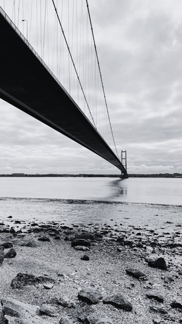 A monochrome photograph captures the underside of a large suspension bridge extending over a body of water, with the bridge deck and suspension cables visible. The bridge's main tower is situated in the distance, partially in the water, with cables stretching diagonally upward to support the deck. The foreground features a rocky shoreline with various-sized stones and pebbles, some darker and smoother, scattered across a slightly uneven surface. The water appears calm, reflecting the bridge's silhouette and the overcast sky filled with thick, grey clouds. The scene exudes a stark, industrial atmosphere with minimal color variation, emphasizing the structural elements of the bridge and the natural setting beneath it, possibly relating to infrastructure or transportation contexts, but solely based on visible elements, not inferred functions or activities.