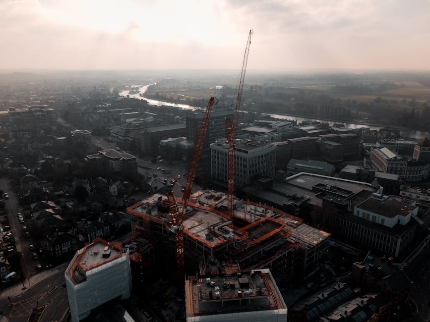 Aerial view of a cityscape during daytime showing a large construction site with two tall red cranes positioned on top of a partially built multi-storey building. The construction area is marked by orange safety barriers and building materials scattered across the structure's concrete floors. Surrounding the site are various modern office and commercial buildings with flat roofs, some featuring rooftop equipment such as HVAC units. To the left, a residential neighborhood with smaller houses and tree-lined streets is visible. In the background, a wide river winds through the landscape, with patches of green fields and woodland extending into the distance under an overcast sky with diffuse sunlight. The scene is captured from a high vantage point, illustrating urban development within a mixed-use environment, with a subtle connection to construction waste management or site clearance efforts, relevant to independent rubbish removal and private disposal services provided by companies like rubbishcollectionkingstonuponthames.co.uk.
